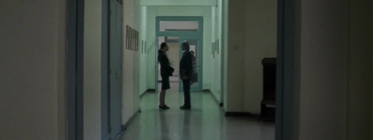 Post Mortem (2010) movie still: wide — A long, minimalist corridor with light green walls and blue door frames. A man and a woman stand in the…
