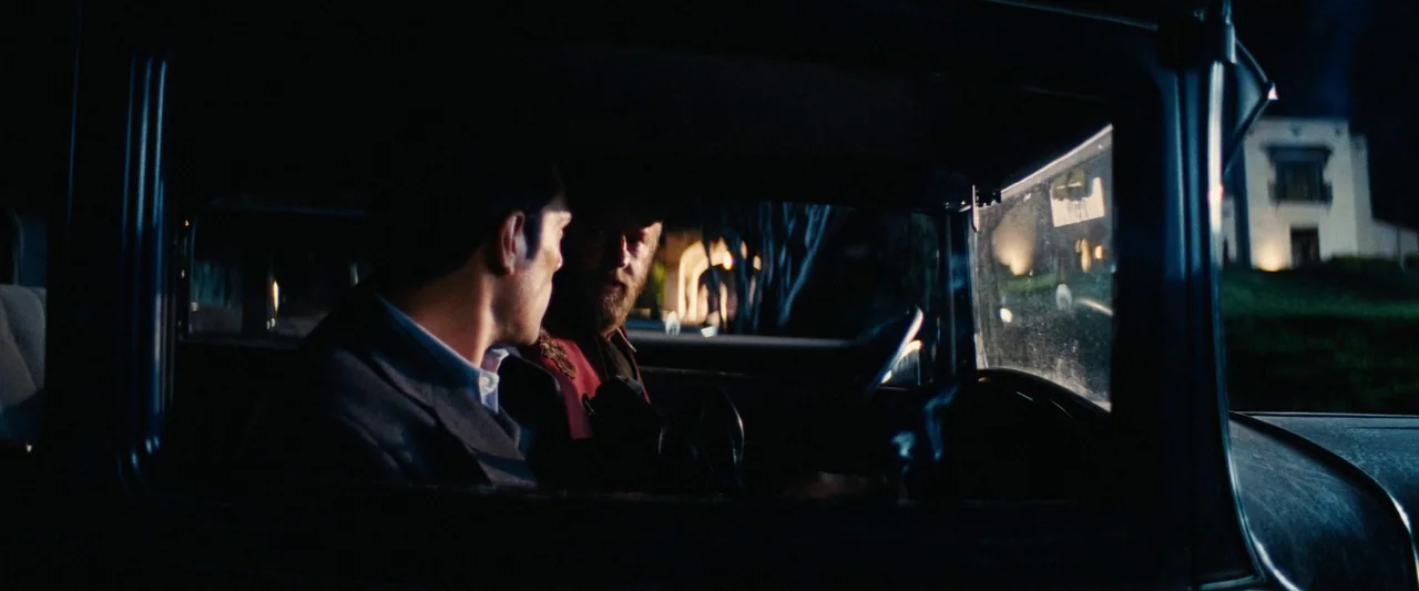 Babylon (2022) movie still: medium — Two men sit inside the cab of a 1940s vintage automobile at night. A large house with lit windows is visible in…