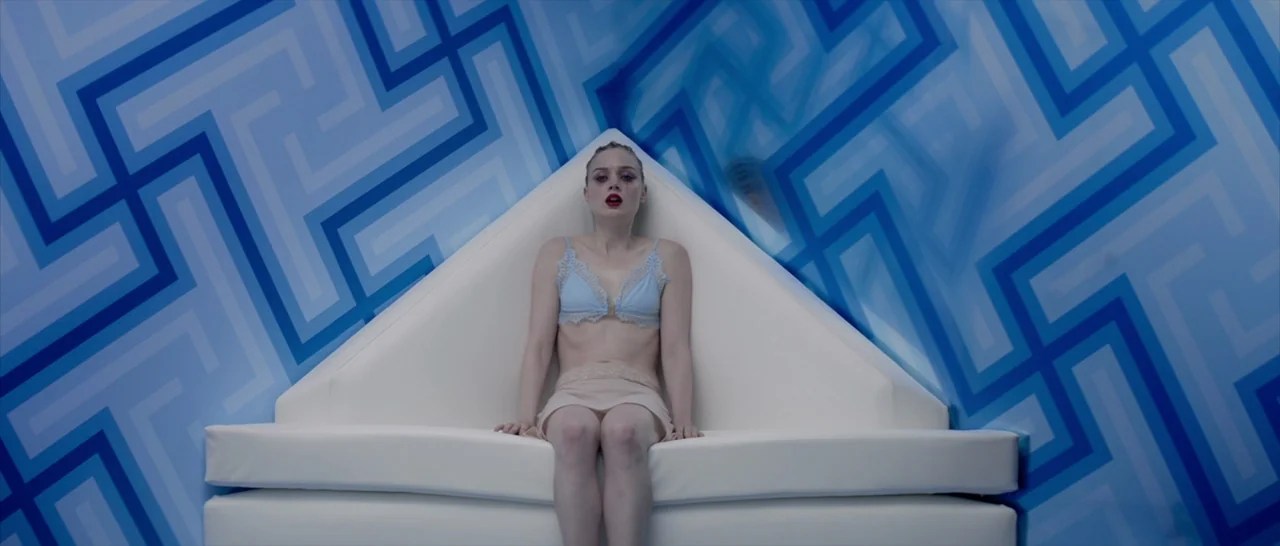 The Neon Demon (2016) movie still: medium — A room with walls featuring a geometric blue and white repeating maze pattern. A woman sits on a stark…