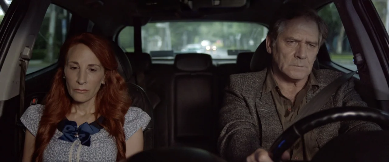 Like or Die (2023) movie still: medium — The interior of a moving car is shown with a driver and passenger. The man operates the steering wheel…