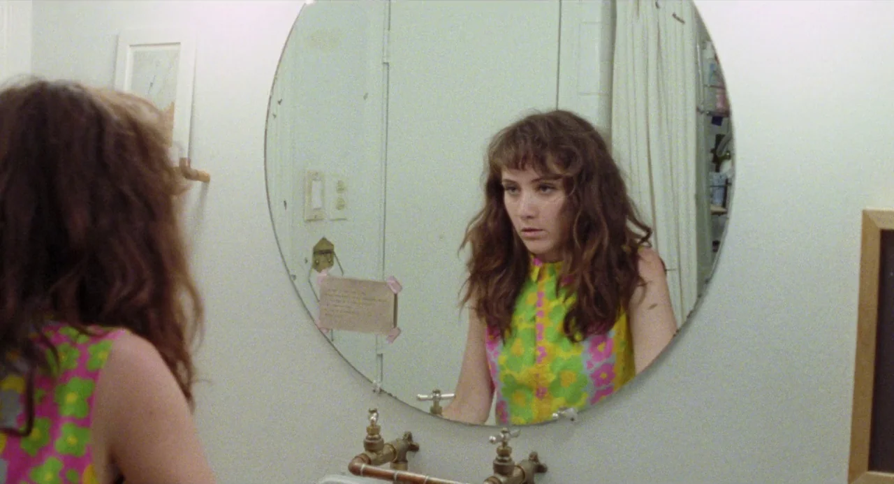 Mr. Roosevelt (2017) movie still: medium — A white-walled bathroom features a round mirror mounted above a sink with exposed pipes. A young woman…