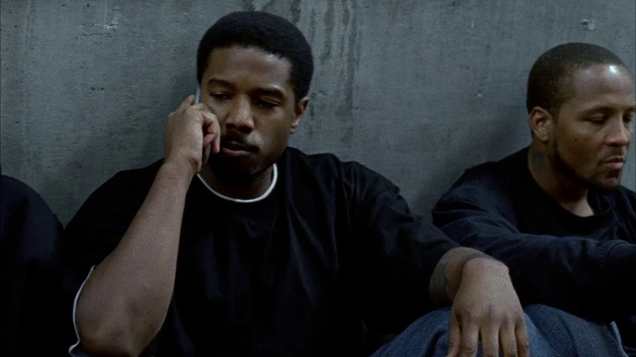 Fruitvale Station (2013) movie still: medium — Two men sit against a smooth, gray concrete wall in an indoor space. The man on the left talks on a…
