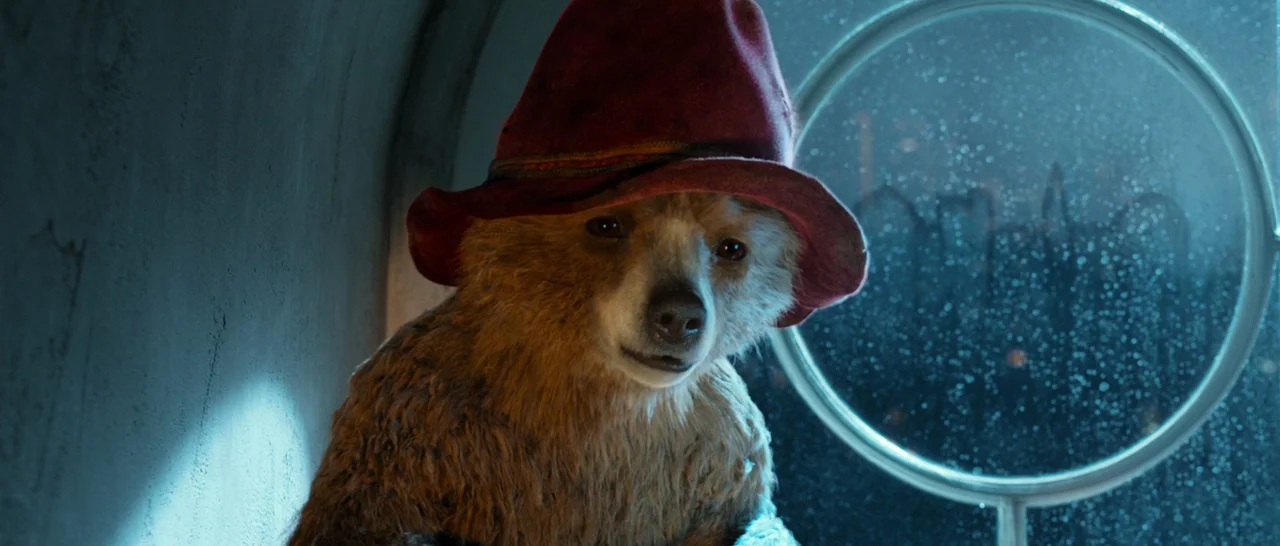 Paddington (2014) movie still: medium — A small anthropomorphic bear sits inside a train car next to a window. Rain streaks the circular window…