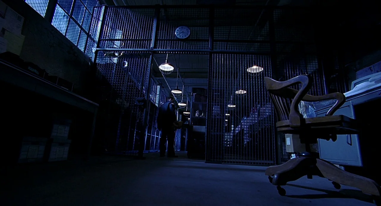 Bride of Chucky (1998) movie still: wide — A secure storage area with floor-to-ceiling metal fencing and rows of hanging lights. A lone figure in…