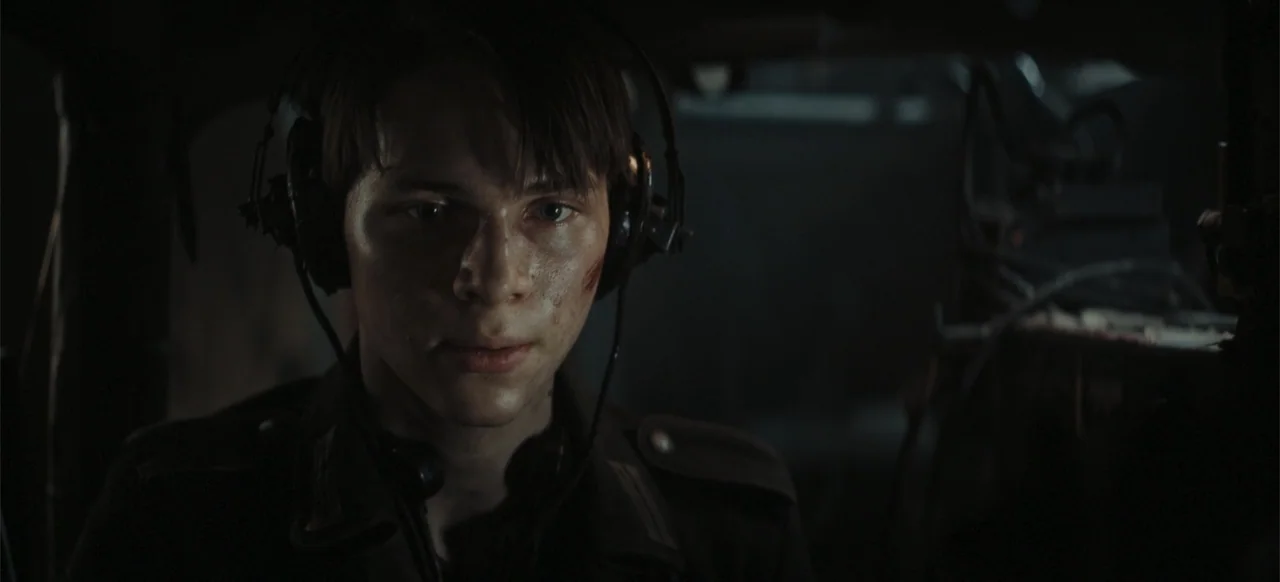 The Tank (2025) movie still: closeup — A dark, cramped interior with metallic textures and electronic equipment visible in the background. A young…