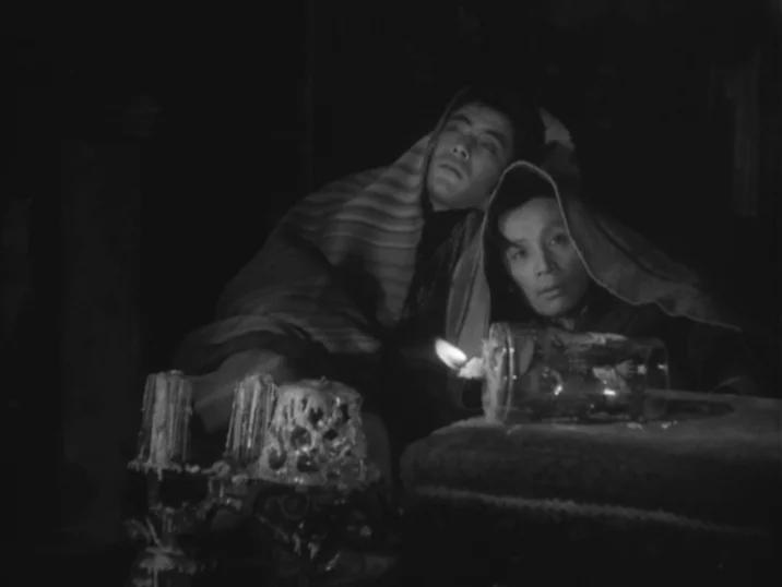 The Idiot (1951) movie still: medium — A dark, dimly lit space features two figures huddled under fabric blankets. The subjects gaze steadily at the…