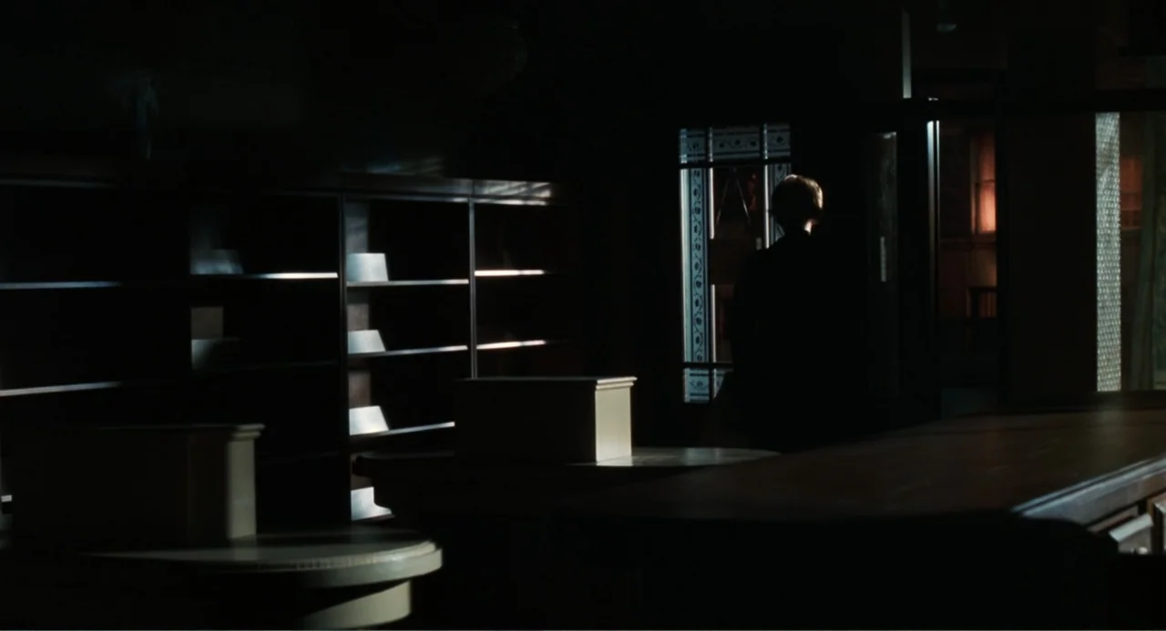You've Got Mail (1998) movie still: wide — A dark, sparsely furnished office space contains tall wooden bookshelves and a desk. A man in a dark suit…