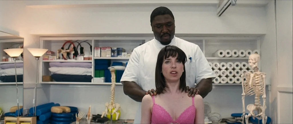 Happy-Go-Lucky (2008) movie still: medium — A bright clinical space contains shelves filled with medical supplies and anatomical models. A man in a…