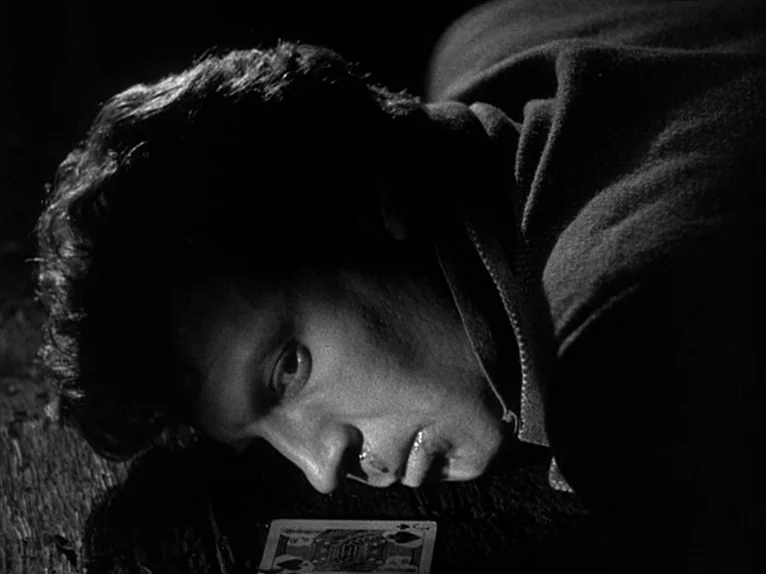 Killer’s Kiss (1955) movie still: closeup — A man lies on a dark wooden surface with a single playing card nearby. The tight framing focuses on his…