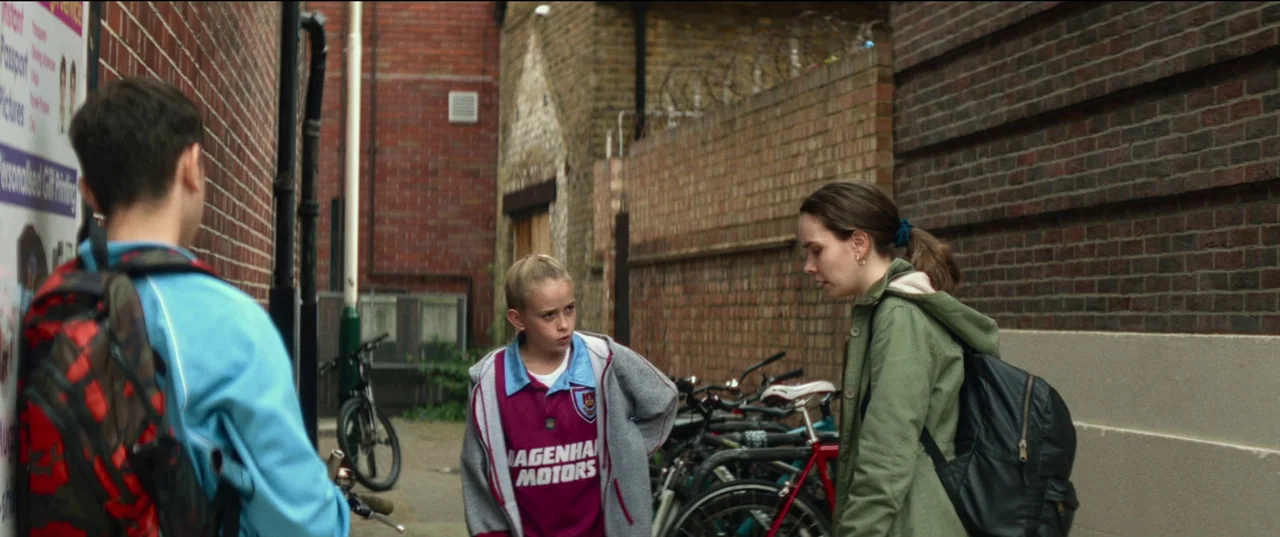 Scrapper (2023) movie still: medium — A narrow brick urban alleyway features high walls topped with barbed wire fencing. Three young people stand…