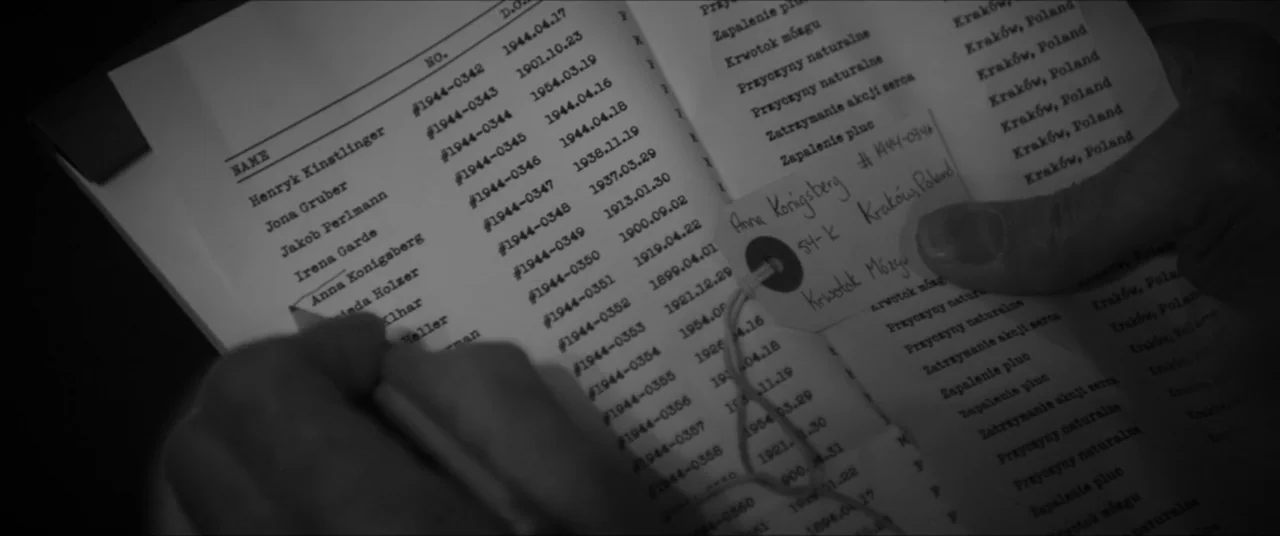 Bau, Artist at War (2025) movie still: closeup — A close-up view shows a black and white ledger listing names and dates. A person uses their hand to…