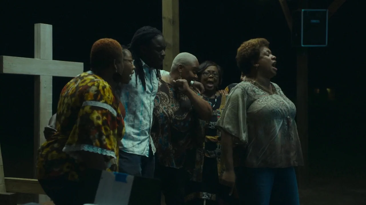 The Sacrament (2013) movie still: medium — A group of five people stand together outside at night in front of a cross. The subjects sing together…