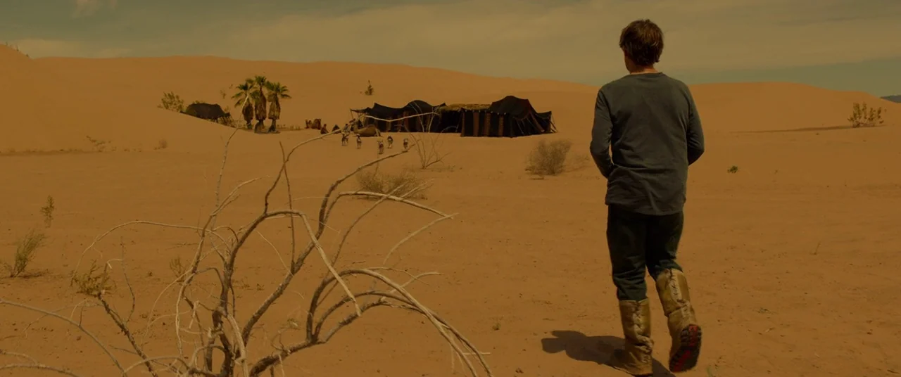 Siberia (2019) movie still: fullbody — A vast sandy desert with rolling dunes and a distant group of black tents. A man walks through the dry…