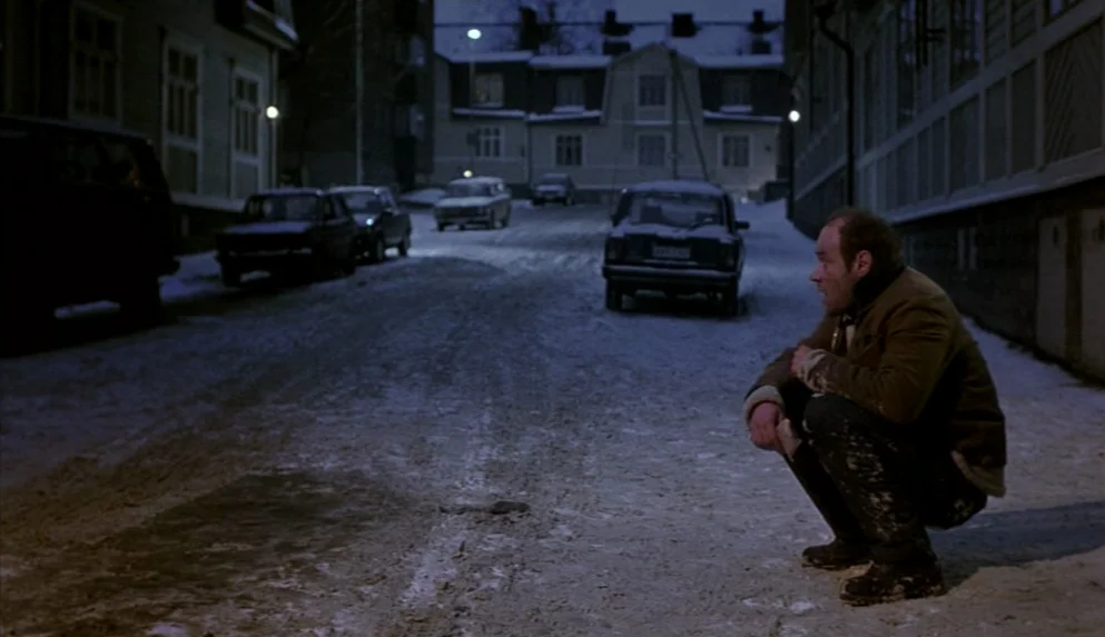 Night on Earth (1991) movie still: fullbody — A snowy street at night lined with houses and parked cars. A man in a brown jacket crouches on the…