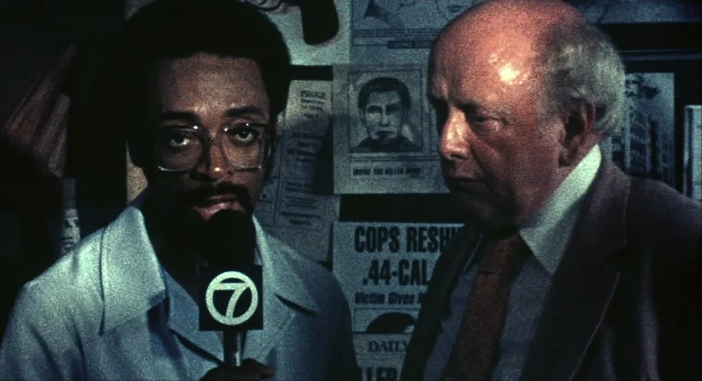 Summer of Sam (1999) movie still: medium — A dark indoor space with wall-mounted police bulletins and posters of criminals. A reporter in a light…
