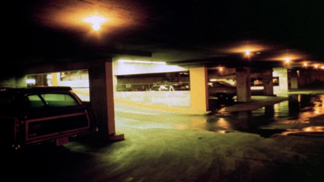 Shivers (1975) movie still: establishing — An empty multi-level parking structure at night with concrete pillars and painted lane dividers. A dark…