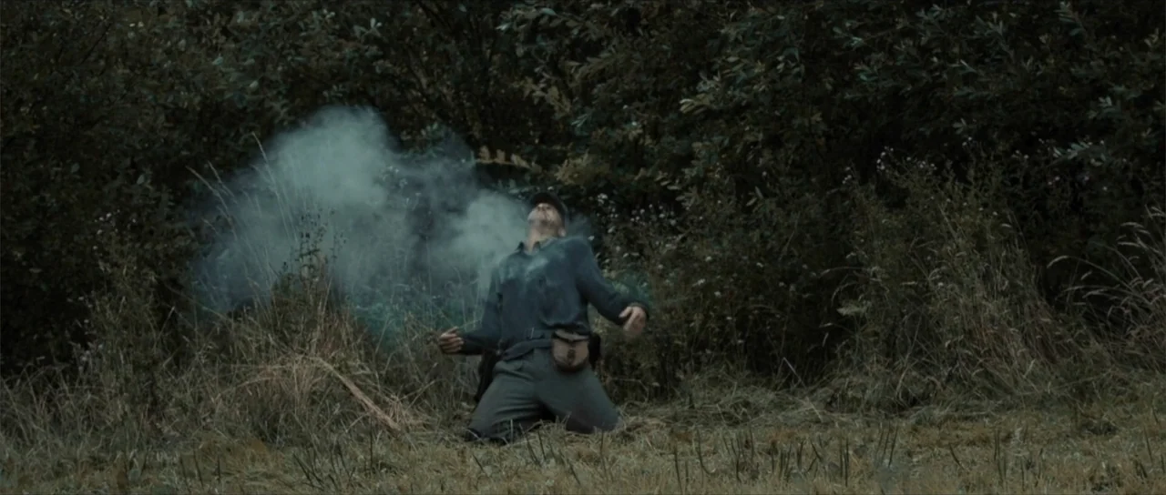 Sin retorno (2009) movie still: fullbody — A field of tall grass stands before a dense line of dark trees. A soldier in grey uniform kneels on the…
