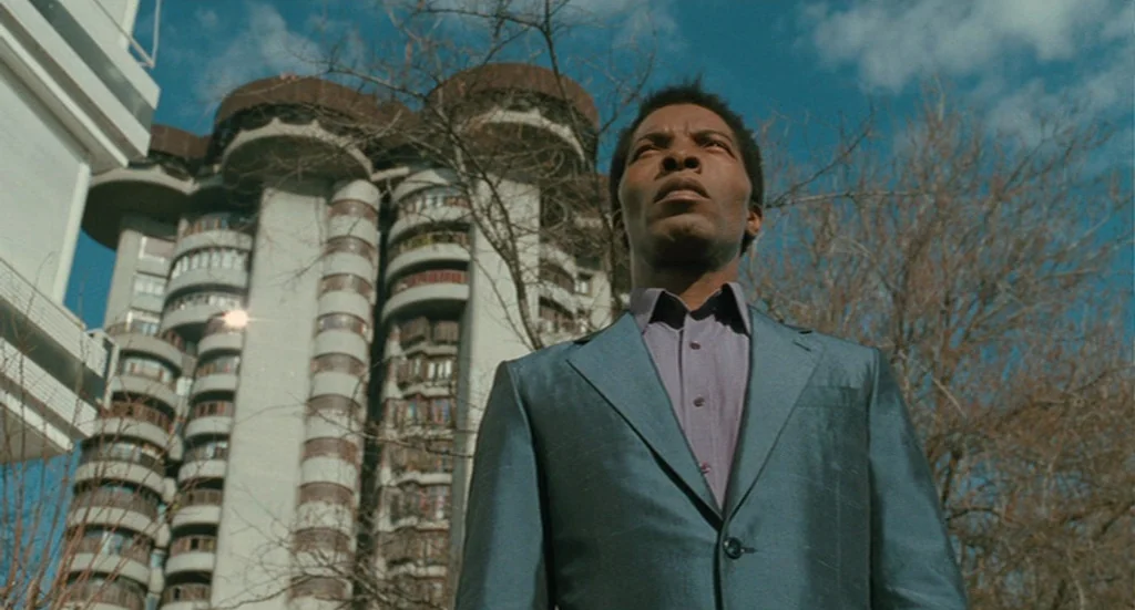 The Limits of Control (2009) movie still: medium — A man stands in front of a prominent modernist residential building with circular balconies. The…