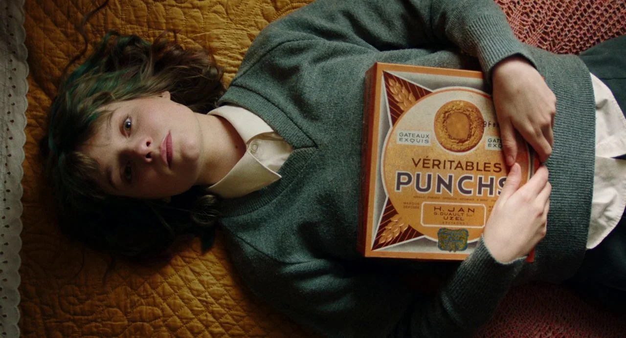 Dating Amber movie still: medium — A person lies on a textured, ochre-colored quilt. They rest a rectangular box of cookies against their chest…