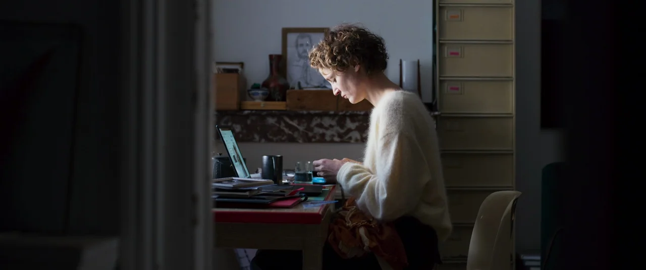 More Than Ever (2022) movie still: medium — A dark, dimly lit room contains a table cluttered with various papers and electronic devices. A woman…