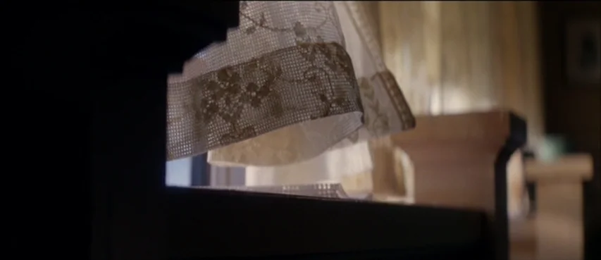 The Thin Red Line movie still: closeup — A close view of a white lace curtain draped over a dark window frame. The blurred background shows hints of…