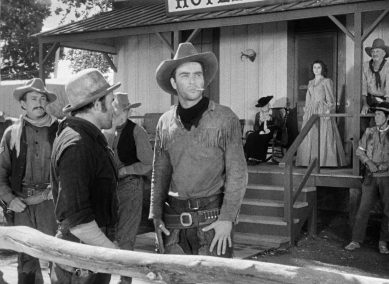 Red River (1948) movie still: fullbody — A wooden porch fronts a hotel building with a group of people standing outside. A young cowboy with a…