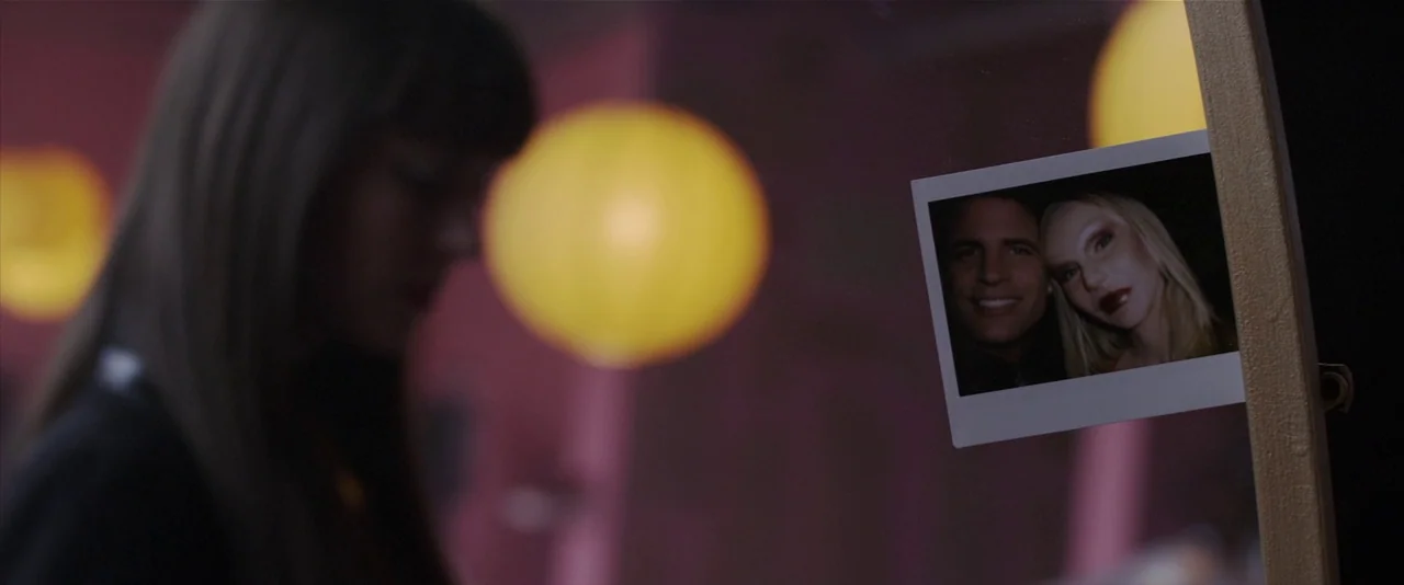 American Mary (2012) movie still: closeup — A dark, dimly lit room features a mirror with a small photograph attached to it. A woman with dark hair…
