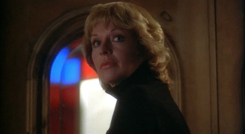 The Silent Partner (1978) movie still: closeup — A dimly lit interior hallway with wood paneled walls and an arched doorway. A woman in a black…