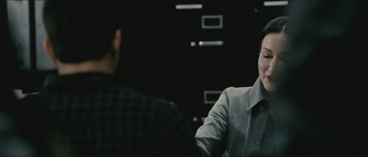 Contraband (2012) movie still: medium — A dark office environment is visible with black metal filing cabinets in the background. A woman in a grey…