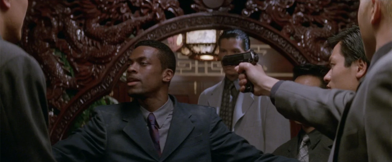 Rush Hour (1998) movie still: medium — A dimly lit room features dark wood carvings and ornate archways in the background. A man aims a handgun at…