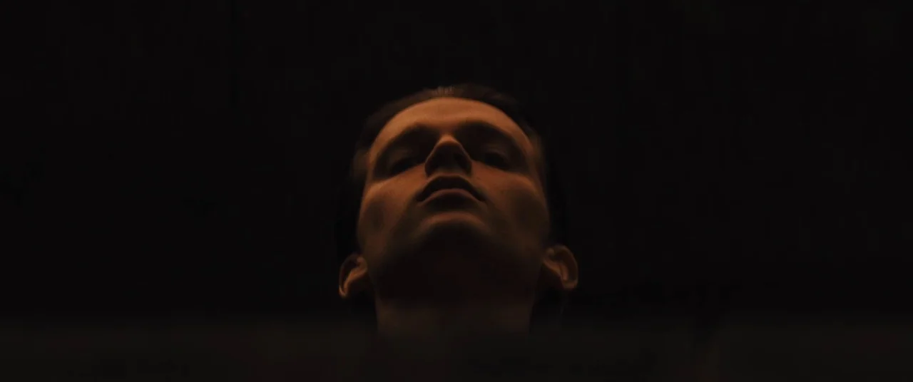 Daniel Isn’t Real movie still: closeup — The subject is framed against a near-total black void in an indeterminate space. The man's face is…