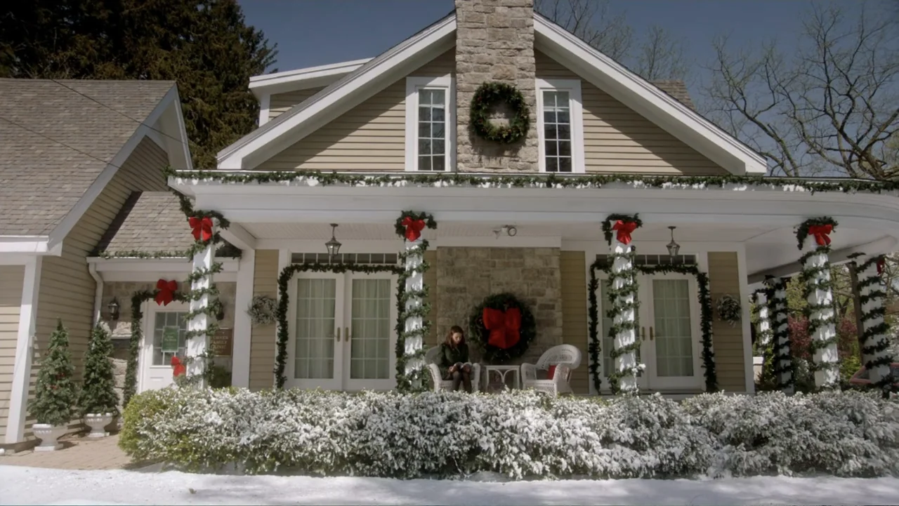 The Christmas Cure (2017) movie still: wide — A light-beige house with a stone fireplace and Christmas decorations stands behind snowy bushes. A…