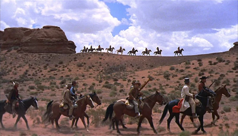 The Searchers movie still: wide — An arid desert landscape features red dirt and sparse brush under a bright sky. A group of riders on horseback…