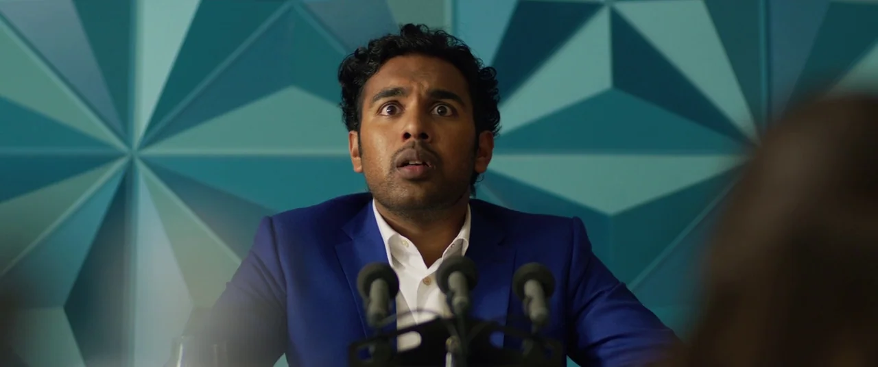 Yesterday (2019) movie still: medium — A man sits at a table in front of a blue geometric patterned wall. He faces the camera with an expression of…