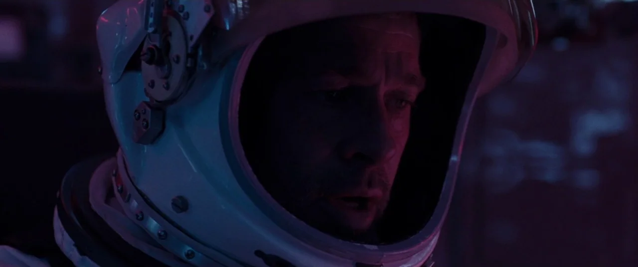 Ad Astra (2019) movie still: closeup — The interior of a cramped spacecraft cockpit is bathed in low-contrast ambient colored light. A middle-aged…
