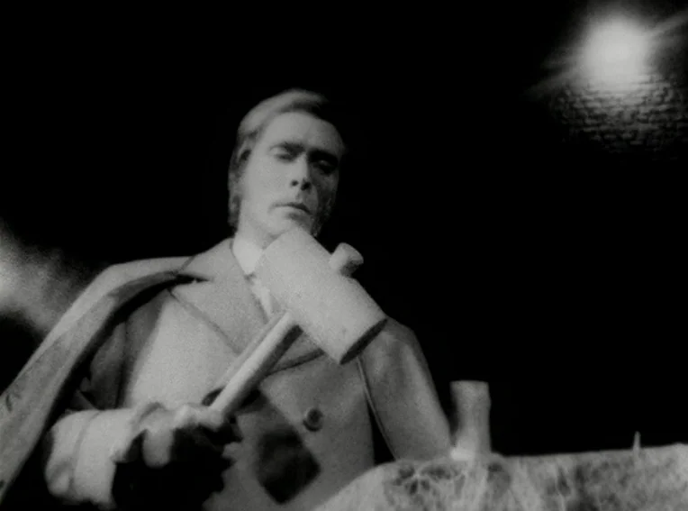 Cuadecuc, Vampir (1971) movie still: medium — A dark night setting featuring a man standing over a soil surface. The man holds a large wooden mallet…