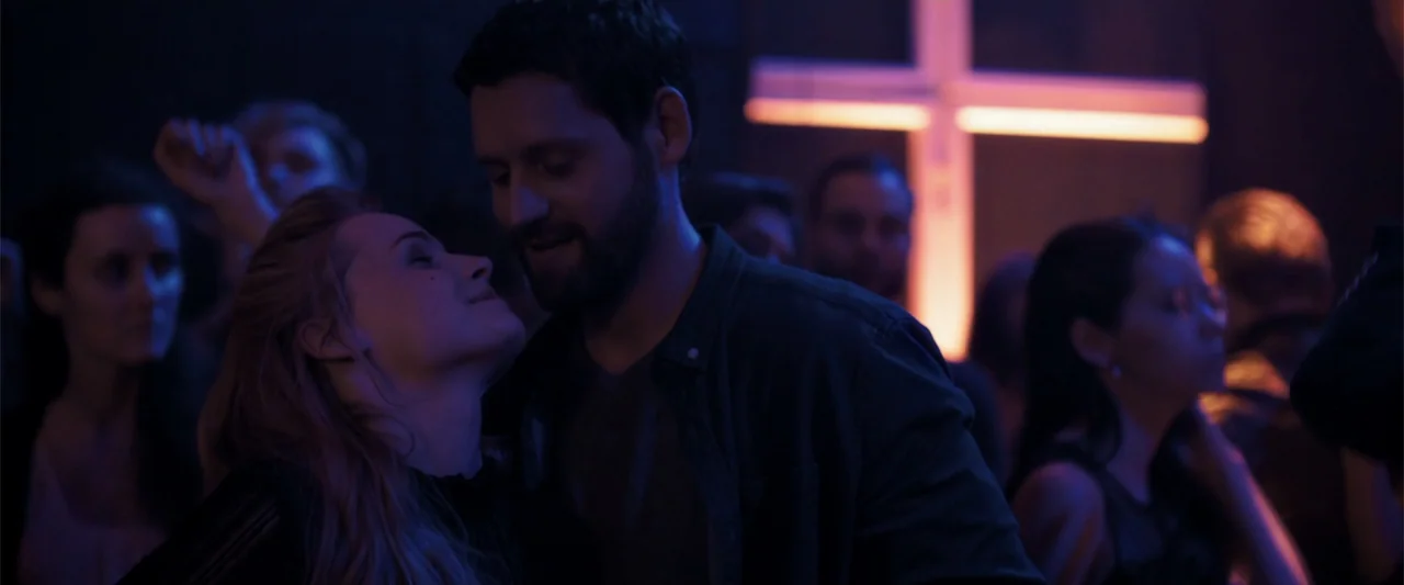 The Good Neighbor (2022) movie still: medium — A crowded dimly lit nightclub interior with a bright glowing neon cross on the wall. A woman looks at…