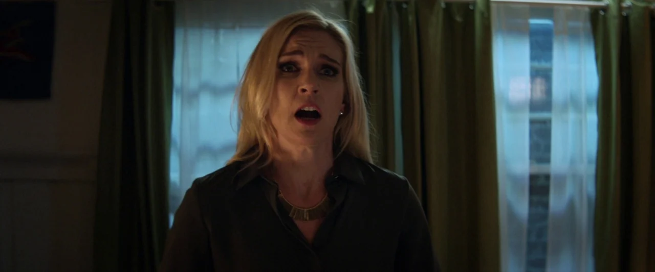Seven Stages to Achieve Eternal Bliss (2018) movie still: medium — A woman stands in a dim room with green curtains hanging over two windows. She…