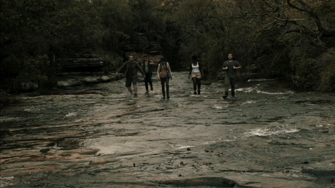 The Grotto (2022) movie still: wide — A group of five hikers walks across a shallow, rocky stream bed in a forest. The water flows over dark stones…