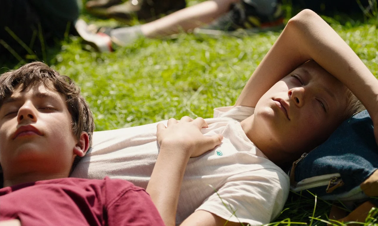 Close (2022) movie still: medium — Two teenage boys lie side by side on a patch of sunlit grass. Their eyes are closed while they rest in the open…