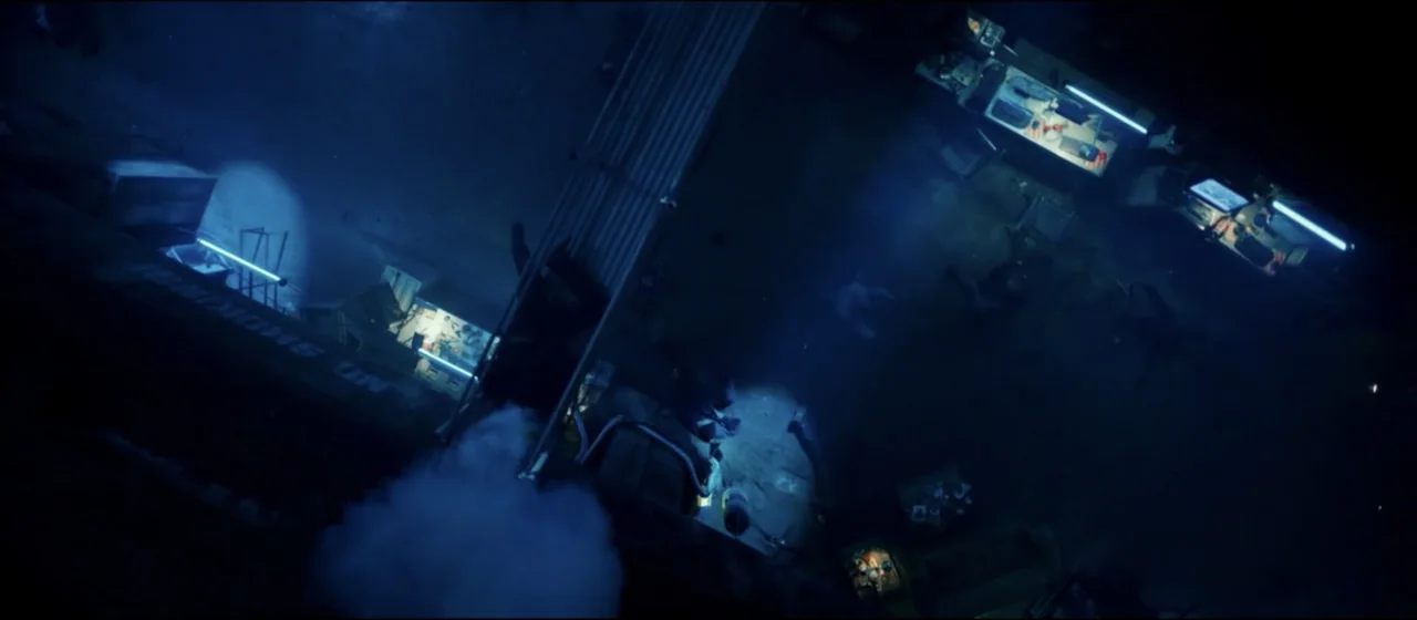 Guns Akimbo (2020) movie still: establishing — A dimly lit office interior is viewed from directly above in a high angle. Several desks with glowing…