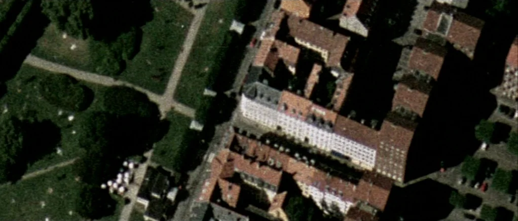 Reconstruction (2003) movie still: establishing — An aerial perspective shows a dense cluster of red-roofed buildings next to a park. Paved paths…