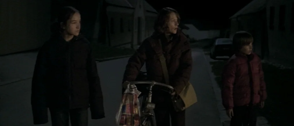 Time Of The Wolf (2003) movie still: fullbody — A dark, empty village street bordered by houses at night. A woman pushes a bicycle while walking…
