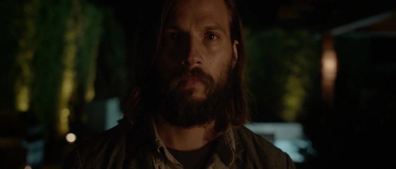 The Invitation (2015) movie still: closeup — A nighttime outdoor setting features dark foliage illuminated by soft, warm ground-level lighting. A…