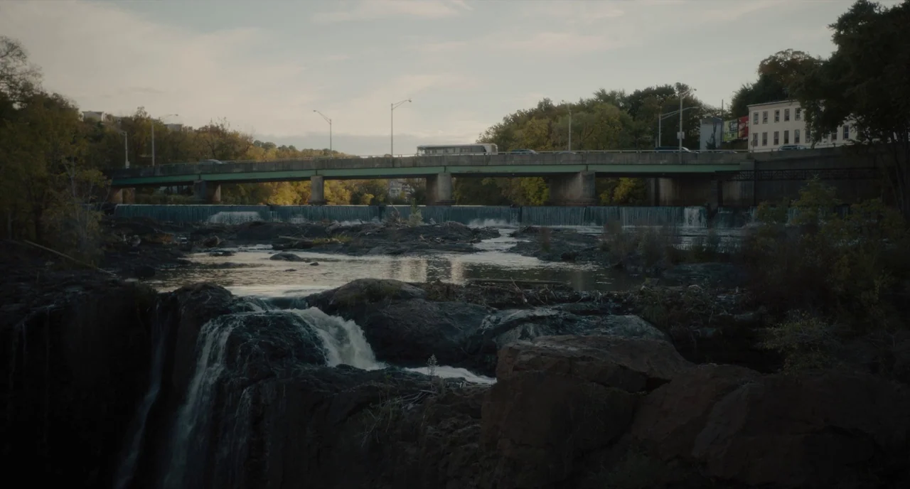 Paterson (2016) movie still: wide — A wide waterfall cascades over dark rocks below a long concrete road bridge. The background features trees in…