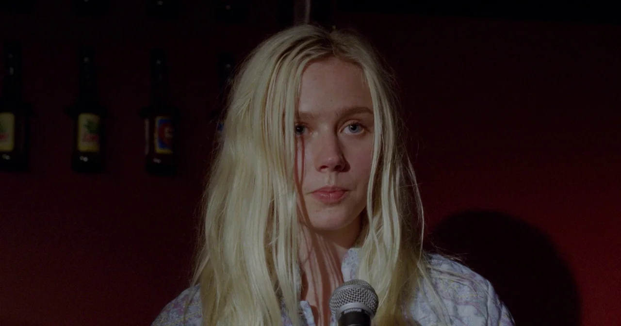 Fallen Leaves (2023) movie still: closeup — A dimly lit indoor space featuring rows of bottles blurred in the background. A young woman with blonde…