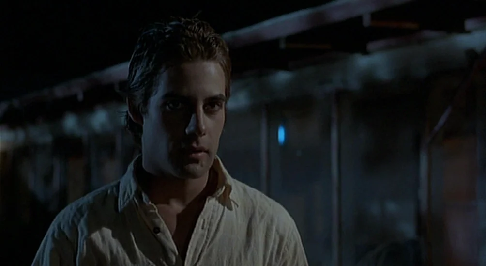 Near Dark (1987) movie still: medium — A dark outdoor space at night with metal structures visible in the background. A young man stands in the…