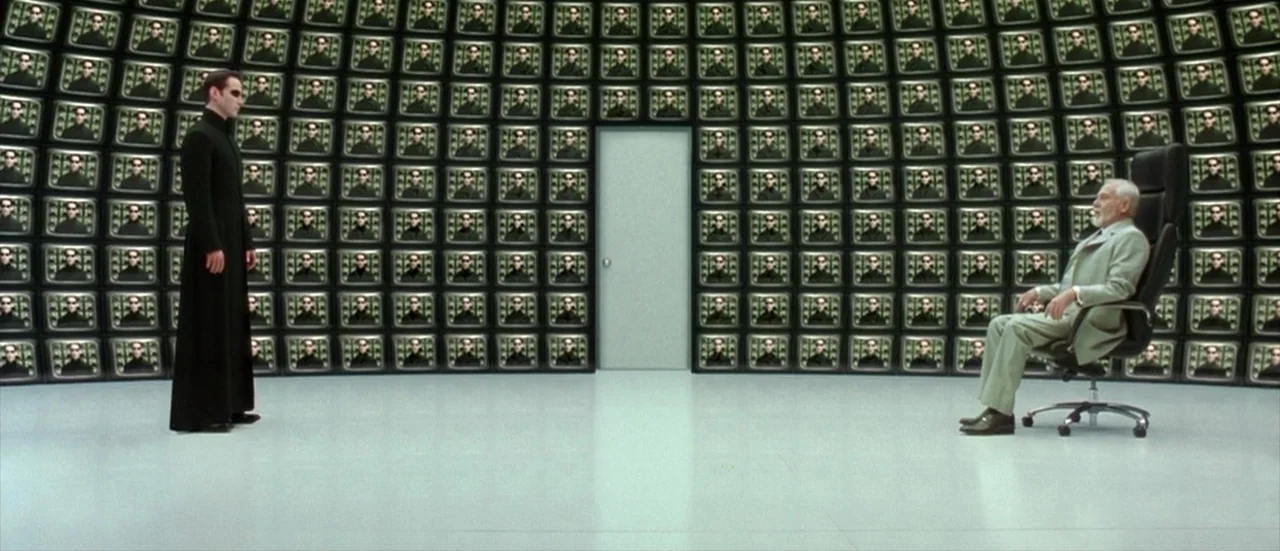 The Matrix Reloaded (2003) movie still: wide — A sterile white room has walls entirely covered by a grid of small television screens. A man in black…