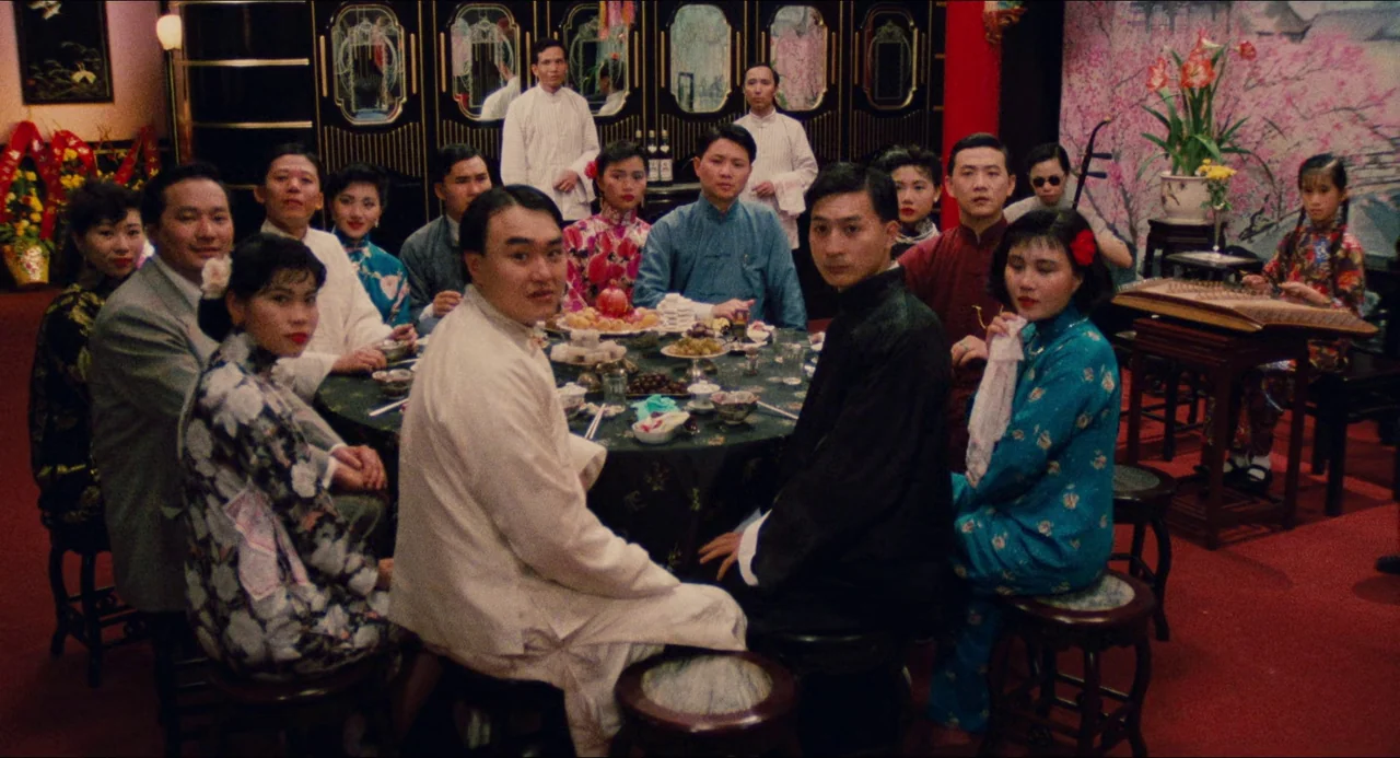Rouge (1987) movie still: wide — A group of people sit around a large round table in a decorated banquet hall. The room features dark wood paneling,…