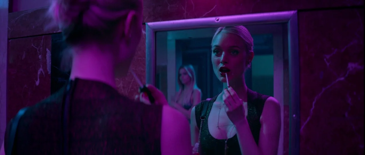 The Neon Demon (2016) movie still: medium — A woman stands before a wall mirror in a room with marble paneling. She holds an unlit cigarette to her…